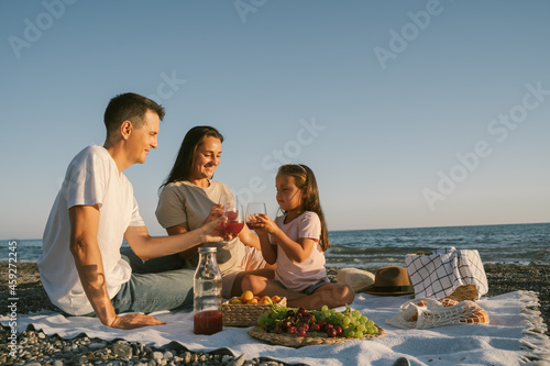 Family spending time together outdoor. Enjoying summer leisure picnic lunch with fruits by the seaside. Happy people eating healthy food and sitting on blanket on the beach.