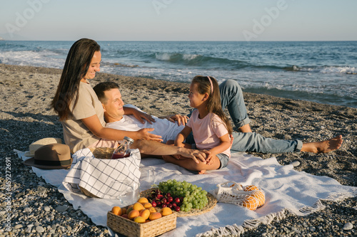 Vegan family spending time together outdoor. Picnic lunch with fruits by the seaside. Happy people eating natural healthy food, drinking juice and sitting on blanket on the beach.