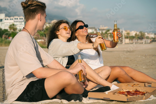 Young happy friends doing picnic seaside. Group of people sitting on the beach, eating pizza and drinking beer together. Summer spending time outdoor concept.