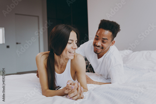 Happy young couple two family in casual white clothes lying in bed on stomach man tickling woman rest relax spend time together in bedroom lounge home house wake up dream be lost in reverie good day. photo