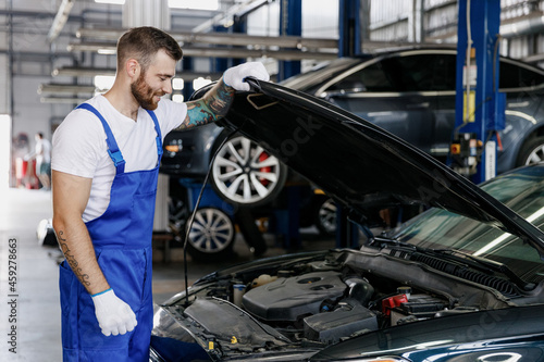 Fun troubleshooter young male professional technician car mechanic man in denim blue overalls white t-shirt fixing problem with raised hood bonnet work in modern vehicle repair shop workshop indoor.