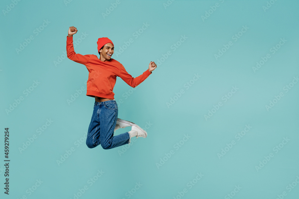 Full body fun cool young smiling happy african american man 20s in orange shirt hat do winner gesture jump high isolated on plain pastel light blue background studio portrait People lifestyle concept