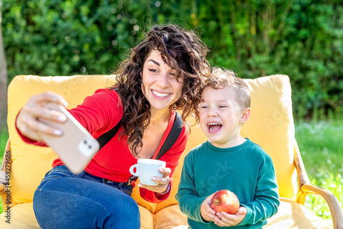 happy mom and son chilling outdoor sitting on a couch in a public park garden making a selfie portrait on smartphone. smiling woman and child interacting online. tech, social media influencer concept