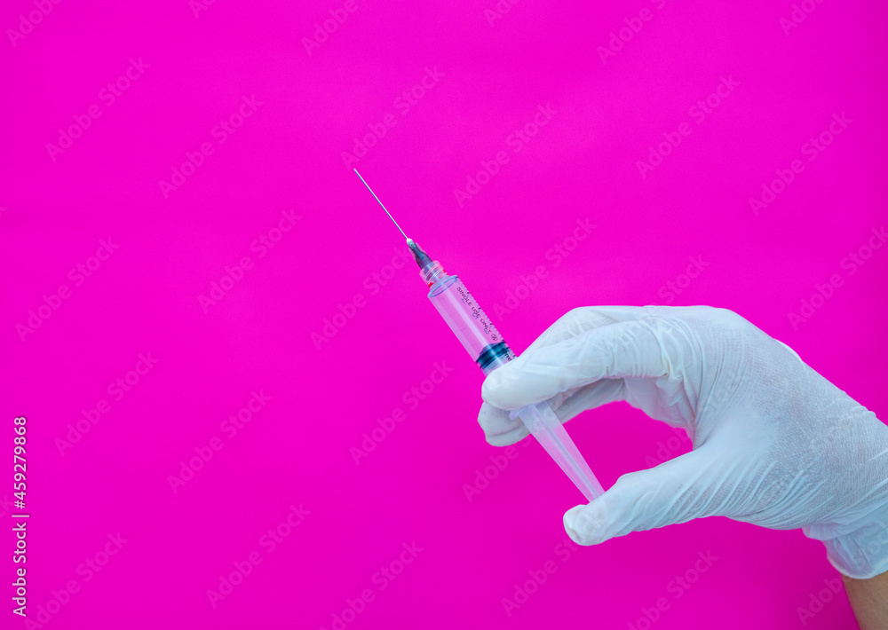 a gloved hand on a pink background, holding a medical syringe that is used to inject drugs and vitamins into the body. a gloved hand on a pink background, holding a medical syringe that is used to inject drugs and vitamins into the body.