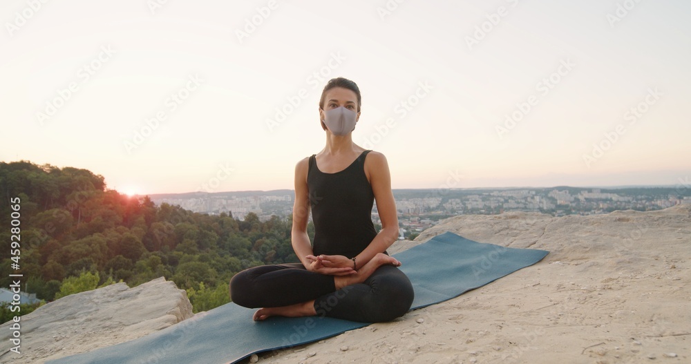 Young caucasian woman wearing protective mask sitting in yoga posture and meditating at the mountains. Girl performing morning meditation at the nature. Physical practice concept
