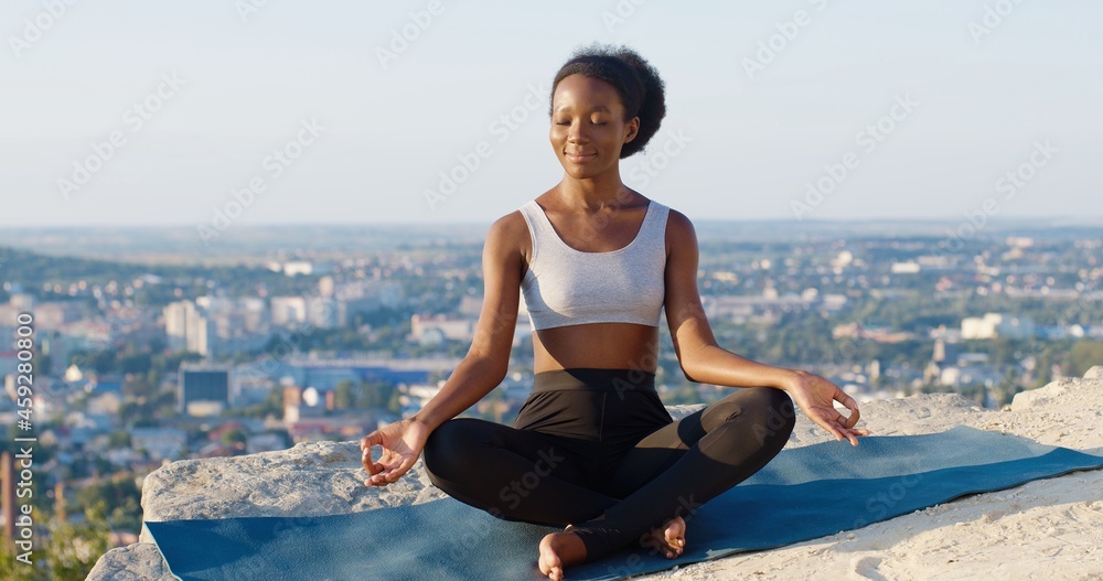 Relaxed calm multiracial woman sitting at the mountain in lotus position, while meditating and practicing yoga with pleasure smile. Enjoying of the morning concept