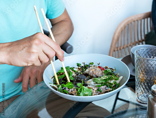 Man in blue t-shirt eating chopsticks healthy vegan food jiaozi with tofu and vegetables in soy sauce with parsley and cilantro in cafe, chinese dish