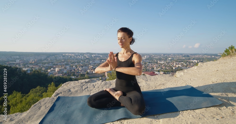 Full length mindful young woman making praying gesture while sitting in lotus position on sports mat at the mountain. Peaceful millennial girl deeply meditating, doing breathing yoga exercises alone