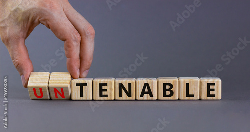 Tenable or untenable symbol. Businessman turns cubes and changes the word 'untenable' to 'tenable'. Beautiful grey table, grey background. Business, tenable or untenable concept, copy space.