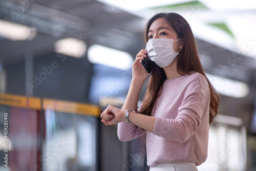 Woman wearing protective mask looks at watch on her wrist at train station platform, waiting someone who is in late, mass rapid transit and crowd at the station in the background