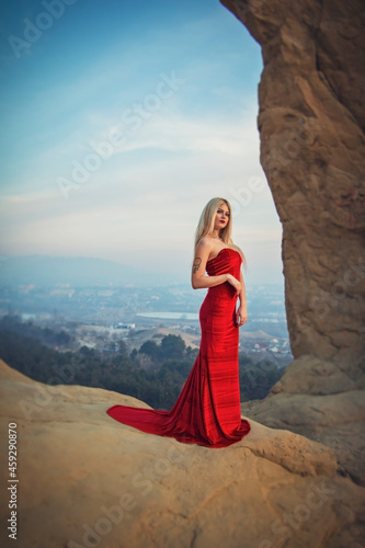 Blonde model girl in a long red dress inside the ring mountain on the background of the city at sunset. On the shoulder is a tattoo of a dream catcher with bird feathers.