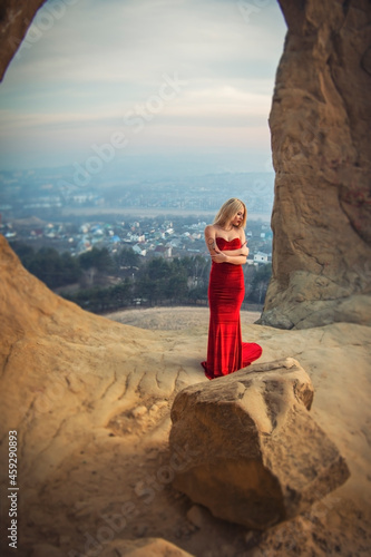 Blonde model girl in a long red dress inside the ring mountain on the background of the city at sunset. On the shoulder is a tattoo of a dream catcher with bird feathers.