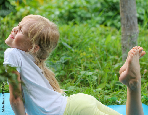 It is difficult for a child to do the exercise, blonde little girl in a white T-shirt does yoga on a blue mat in the garden. a child and a healthy lifestyle