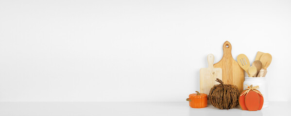 Autumn theme kitchenware, utensils and pumpkin decor on a white shelf or counter. Banner against a white wall background with copy space. Fall home kitchen cooking decor.