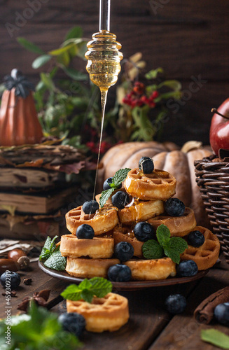 many waffles are stacked on a plate, decorated with mint leaves and blueberries. On top of them, honey pours from glass spoons