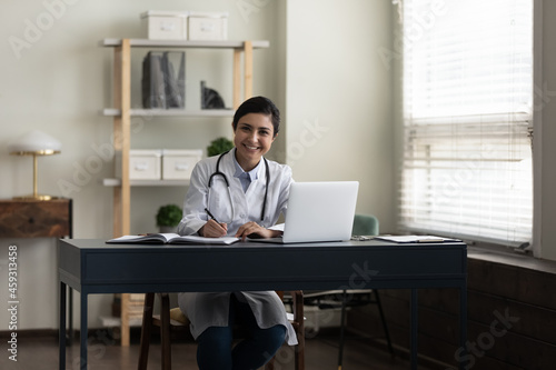 Happy young Indian doctor, therapist, general practitioner at work table with laptop in office, looking at camera, smiling, writing notes, medical records. Medic care concept. Professional portrait © fizkes