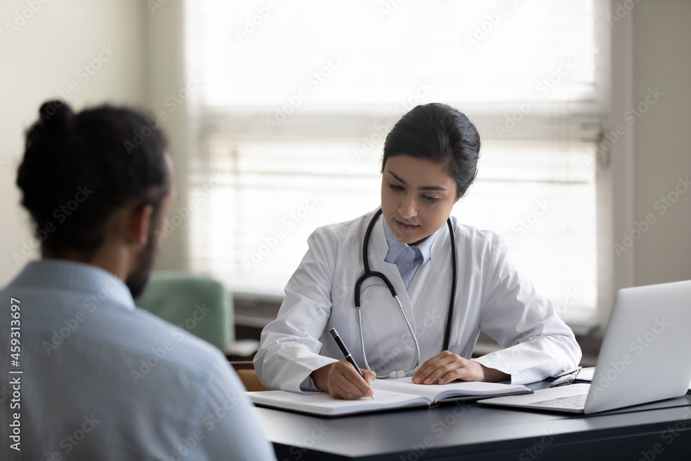Serious Indian GP doctor giving consultation to patient in hospital office, listening to complaints, writing medical records in register. Young mixed race man visiting, consulting therapist Serious Indian GP doctor giving consultation to patient in hospital office, listening to complaints, writing medical records in register. Young mixed race man visiting, consulting therapist