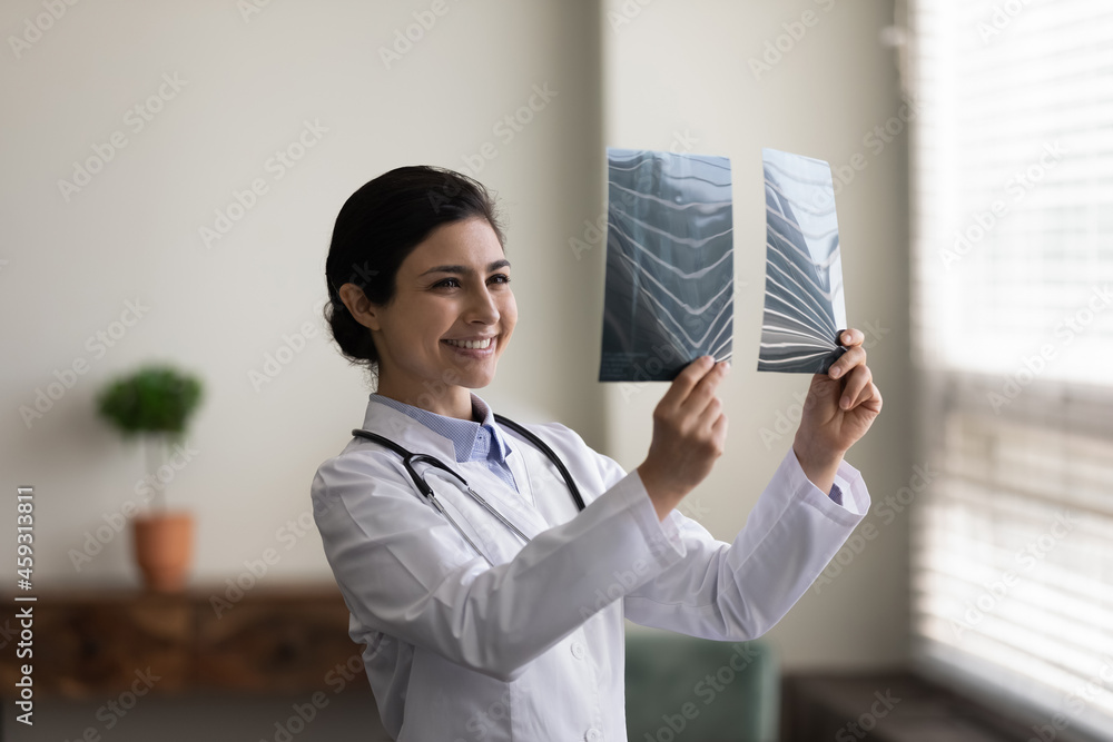 Happy young Indian doctor looking at xray radiography images. Physician, surgeon reviewing scan of patient bones, smiling at good screening test result. Medical checkup, healthcare, radiology concept Happy young Indian doctor looking at xray radiography images. Physician, surgeon reviewing scan of patient bones, smiling at good screening test result. Medical checkup, healthcare, radiology concept