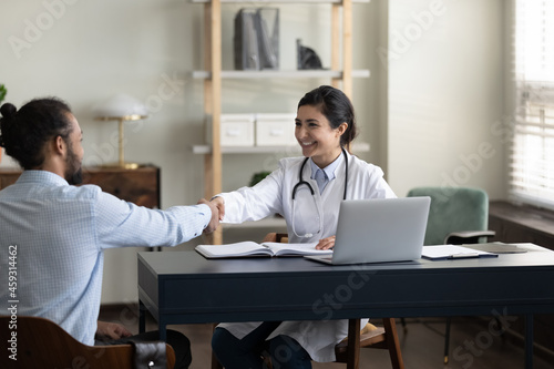 Happy young Indian GP doctor shaking hands with male patient, celebrating successful therapy result. Mixed raced millennial man visiting practitioner, shaking hands, expressing gratitude, respect