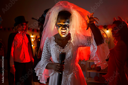 Portrait of black woman in white bridal veil at Halloween horror party at home. Escape room actress who plays Corpse Bride dead girl holding flashlight that lights up face with ugly grimace expression photo
