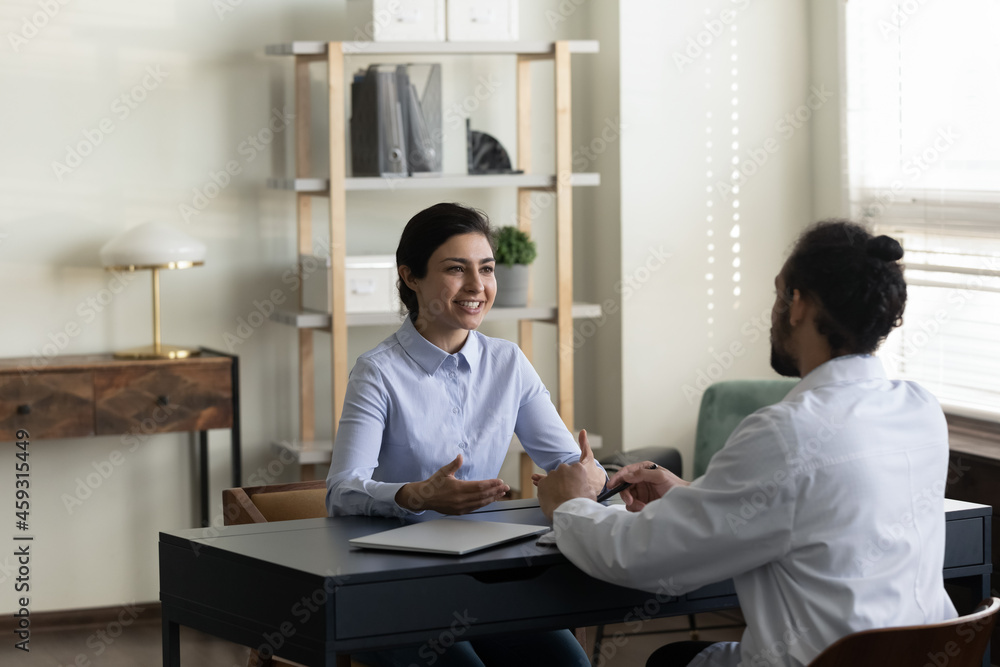Obraz premium Happy Indian woman consulting young male doctor at appointment. Millennial mixed race general practitioner meeting with patient, listening complaints, giving treatment advice, prescription