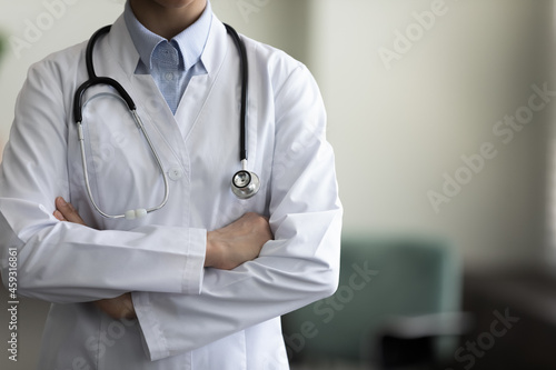 Cropped shot of female GP doctor with stethoscope wearing white coat, standing indoors with folded arms. General practitioner, physician, medical expert in office close up portrait