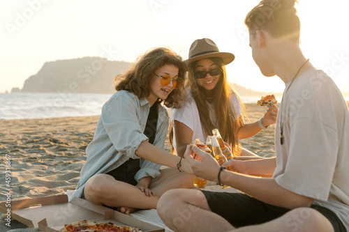 Young happy friends doing picnic seaside. Group of people sitting on the beach, eating pizza and drinking beer together. Summer spending time outdoor concept. © valeriyakozoriz