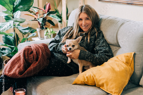 Young woman with plaid and pillow sitting on couch at home  wearing comfortable clothes and playing with her chihuahua dog. Autumn fall and winter season. Lifestyle photography