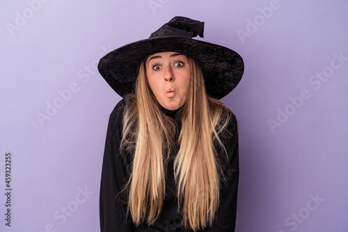 Young Russian woman disguised as a witch celebrating Halloween isolated on purple background shrugs shoulders and open eyes confused.