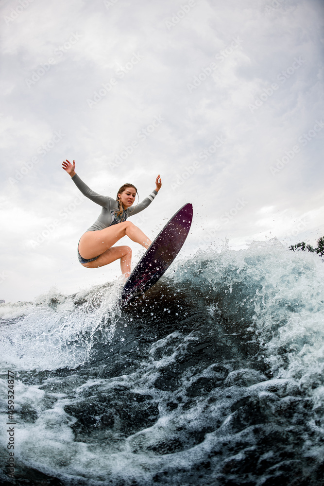 Attractive athletic woman in grey swimsuit on surfboard jumps at river wave