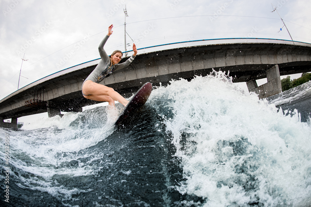 Naklejka premium energetic woman rides down the wave on wakesurf board. Bridge at background