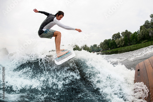 Athletic man active riding wave on surf style wakeboard and lifting up lot of splashes