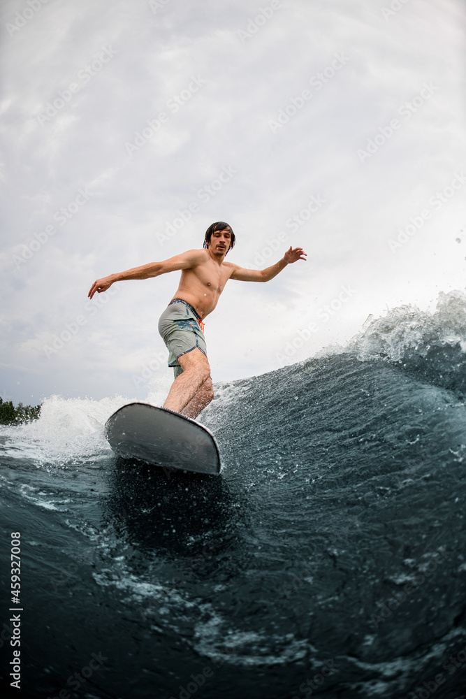 athletic man stands on a wakesurf board and rides down on river wave on summer day
