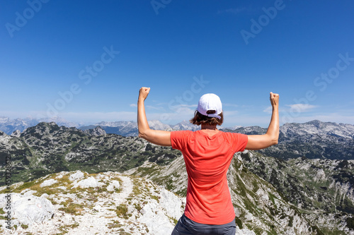Woman celebrating mountaineering achievement with arms raised in the air, while looking at beautiful mountaintops landscape