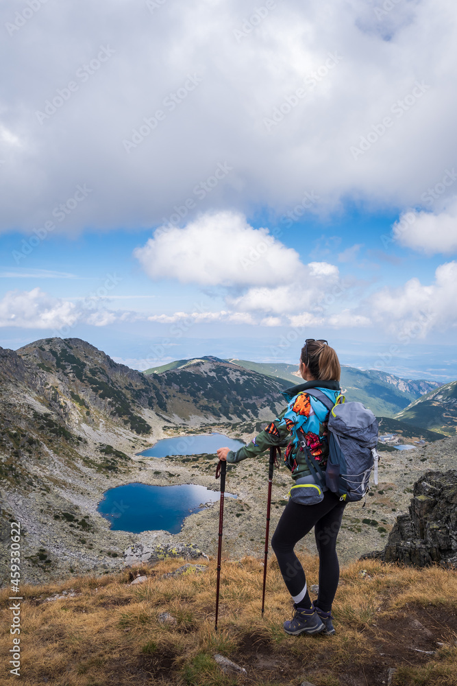 Girl mountain hiker in colorful clothes and with walking sticks standing on a cliff and looking at beautiful Glacier lakes on Rila mountain