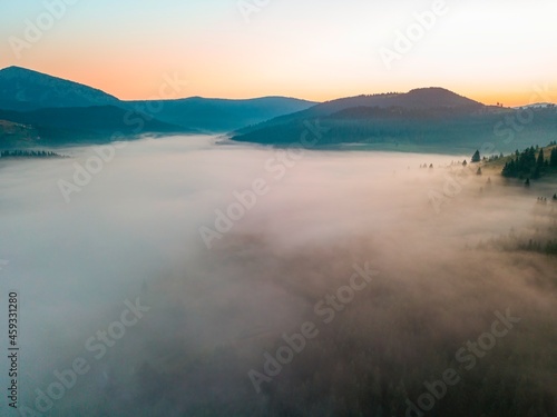 Fog spreads over the mountains at dawn. The sun rises on the horizon. Ukrainian Carpathians in the morning. Aerial drone view.