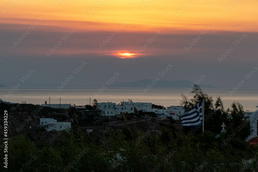 Sun Setting in the Distance Along the Water in Mykonos by a Greek Flag Flying