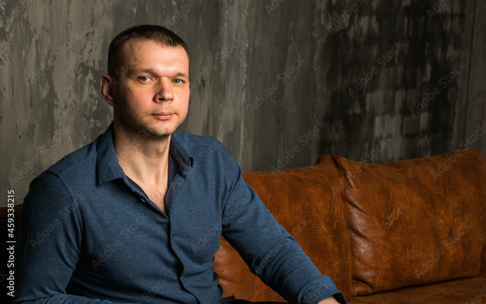 A young man in a blue shirt is sitting on a vintage sofa in a dark room, against a brick wall. A young man in a blue shirt is sitting on a vintage sofa in a dark room, against a brick wall.