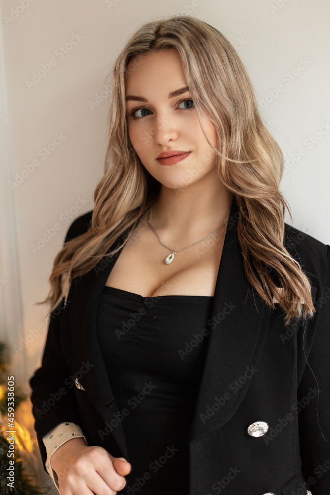 Cute beautiful young woman in fashionable black clothes with an elegant blazer and big breasts stands near a white wall Cute beautiful young woman in fashionable black clothes with an elegant blazer and big breasts stands near a white wall