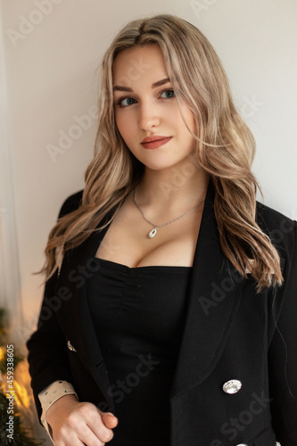 Cute beautiful young woman in fashionable black clothes with an elegant blazer and big breasts stands near a white wall