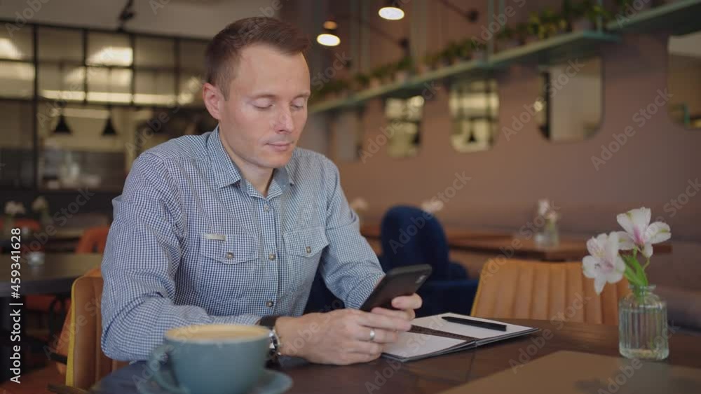 Medium shot of young man wearing streetwear sitting in restaurant or cafe in front of laptop and having mobile phone. Breakfast With Smartphone Medium shot of young man wearing streetwear sitting in restaurant or cafe in front of laptop and having mobile phone. Breakfast With Smartphone
