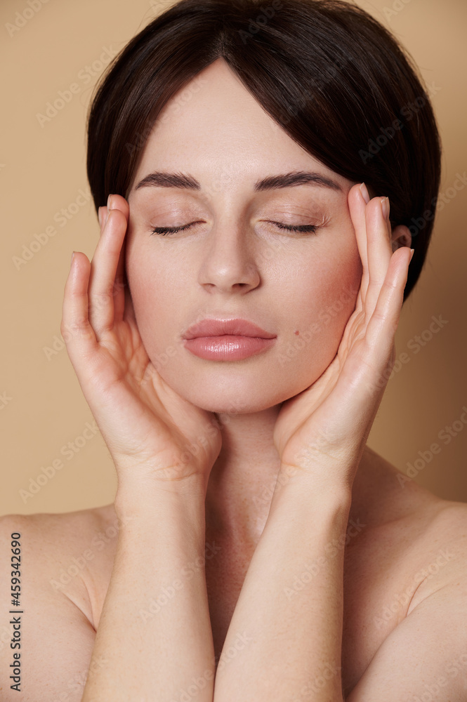 Close-up portrait of a half naked beautiful woman with natural make-up and perfect clean shiny skin massaging her temples, posing with closed eyes against beige background with copy space. SPA concept