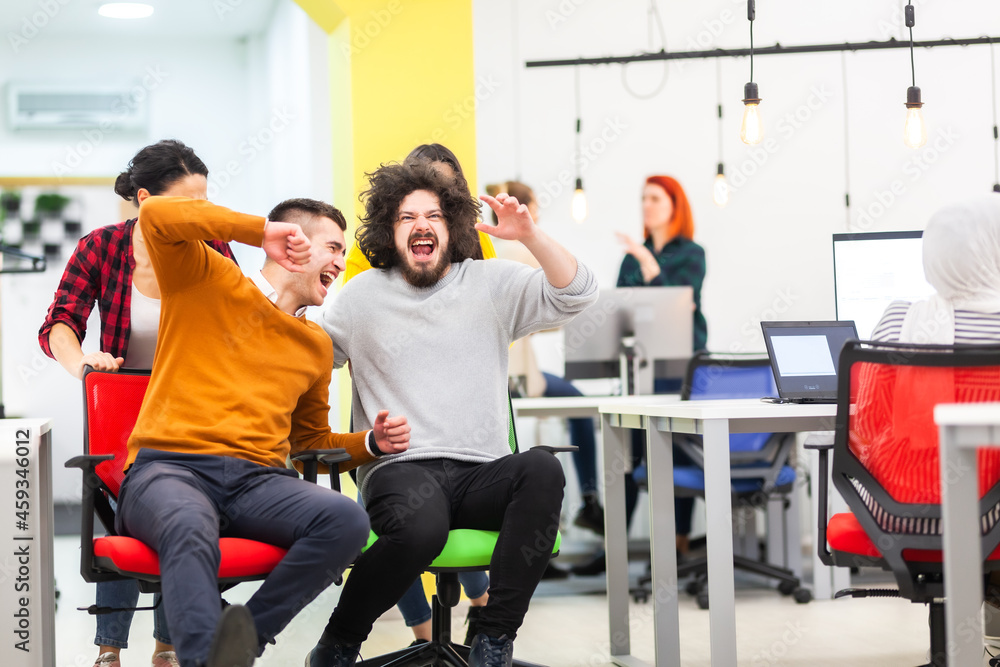 Colleagues having fun on work at the office. Girls pushing boys on chairs with wheels. Multiethnic group of successful start up company. Colleagues having fun on work at the office. Girls pushing boys on chairs with wheels. Multiethnic group of successful start up company.