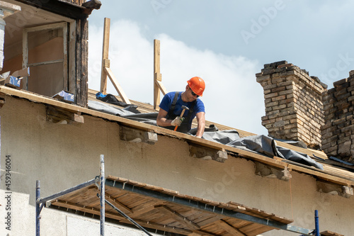 Repair of a wooden roof outdoors on a summer day against the background of blue sky and clouds. A carpenter in special clothes and with a tool installs beams and wood boards.