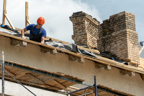 Repair of a wooden roof outdoors on a summer day against the background of blue sky and clouds. A carpenter in special clothes and with a tool installs beams and wood boards.