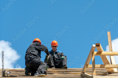 Repair of a wooden roof outdoors on a summer day against the background of blue sky and clouds. A carpenter in special clothes and with a tool installs beams and wood boards. © Pokoman