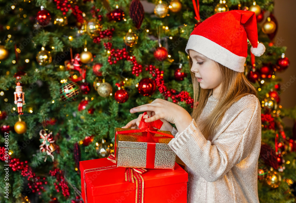Happy beautiful girl in a Santa hat looks at the gifts and presents at the Christmas tree with a smile Happy beautiful girl in a Santa hat looks at the gifts and presents at the Christmas tree with a smile