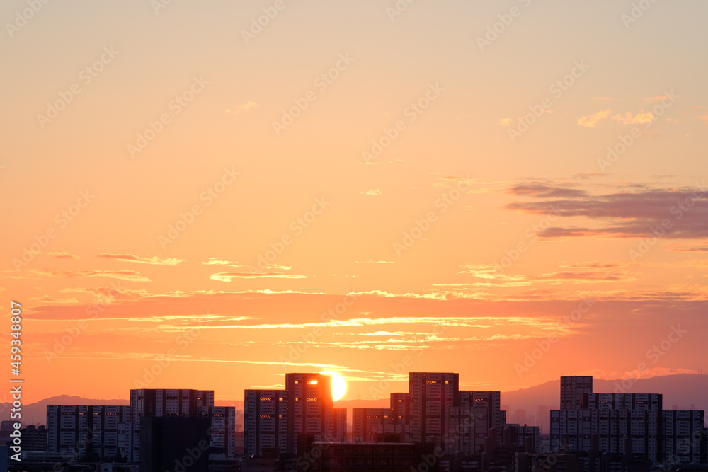 都市の夜明け、早朝ビルの隙間から太陽が昇り辺りはオレンジ色に染まる。ビルはシルエットに浮かぶ。