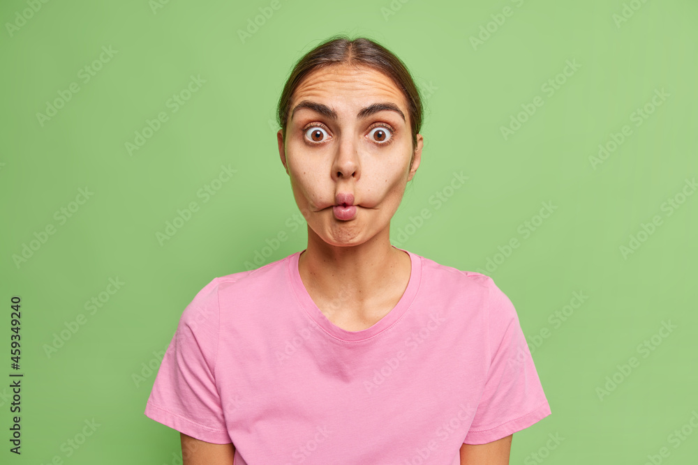 Fototapeta premium Surprised young woman makes fish lips being not scared to show emotions tries to make her crying friend laugh dressed in casual pink t shirt isolated over green background. Face expressions.