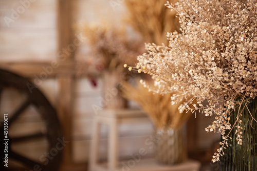 In green glass vase, there was bouquet of dried flowers on wooden bench near backdrop of rural style